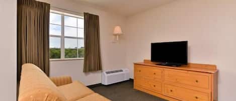 Una habitación de hotel con sofá, cómoda de madera, televisor y ventana con cortinas.