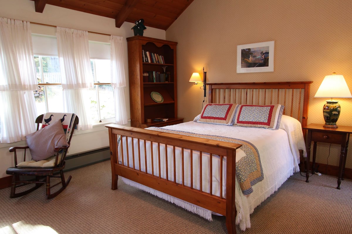 A bedroom with a wooden bed, a rocking chair, a bookshelf, and a lamp.