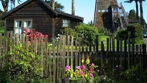 A wooden cabin with a stone tower, surrounded by a wooden fence and lush greenery. - Joshua Grindle Inn (Mendocino)