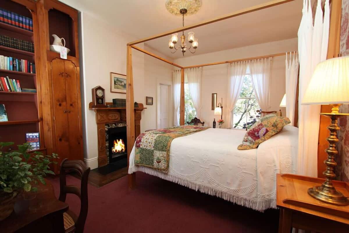 A bedroom with a four-poster bed, a fireplace, a bookshelf, and a window with curtains.