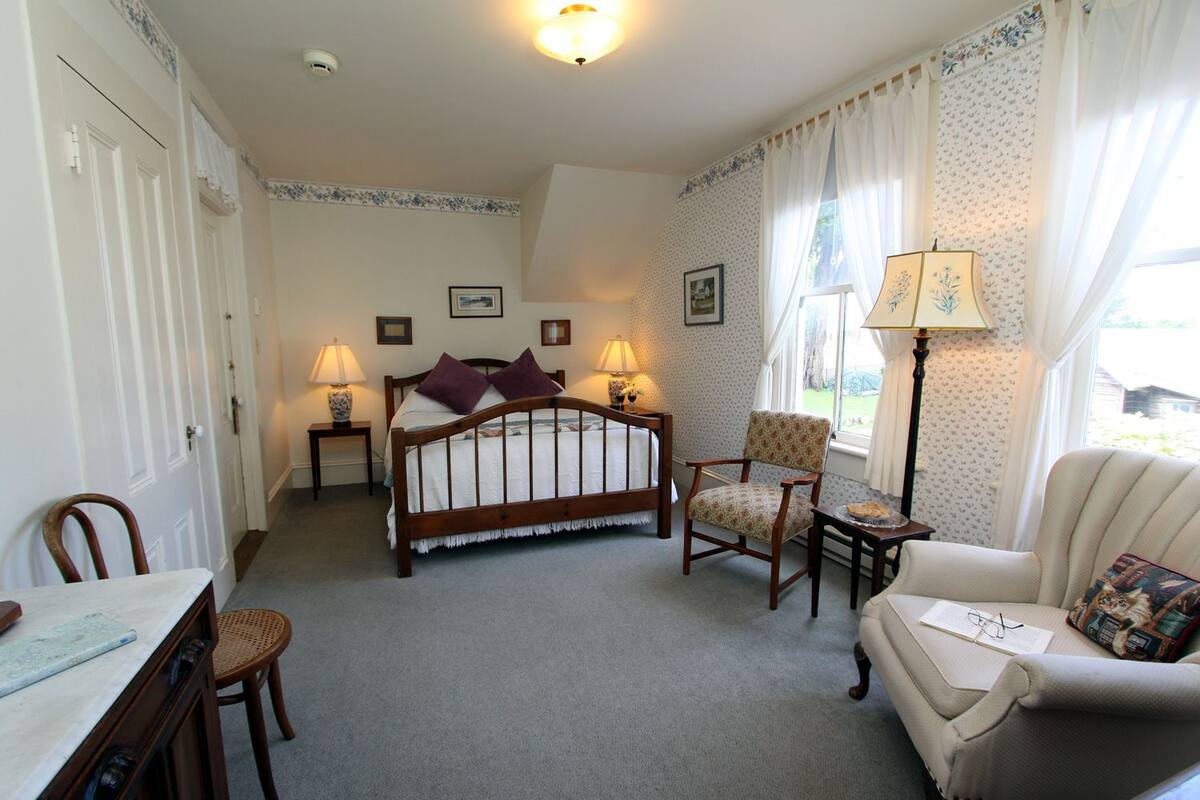 A bedroom with a wooden bed, two bedside tables with lamps, a chair, a sofa, and a small table with a book and glasses.