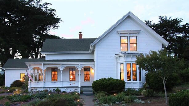 A white two-story house with a porch and bay windows, surrounded by a garden with various plants and a small American flag. - Joshua Grindle Inn (Mendocino)
