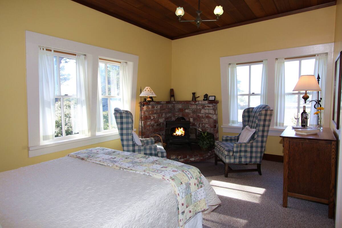 A cozy room with a bed, two plaid chairs, a fireplace, and a lamp on a wooden table.