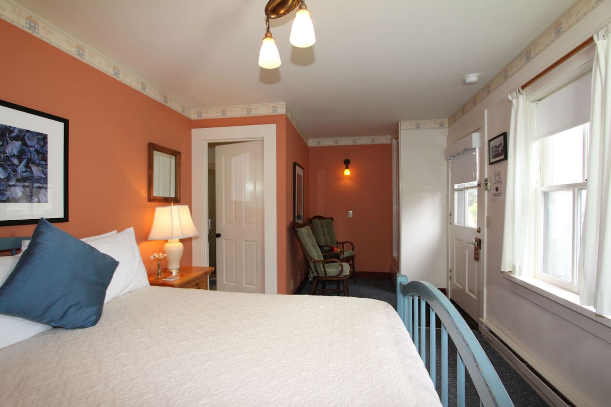 A hotel room with a bed, a chair, a lamp, a door, and a window.