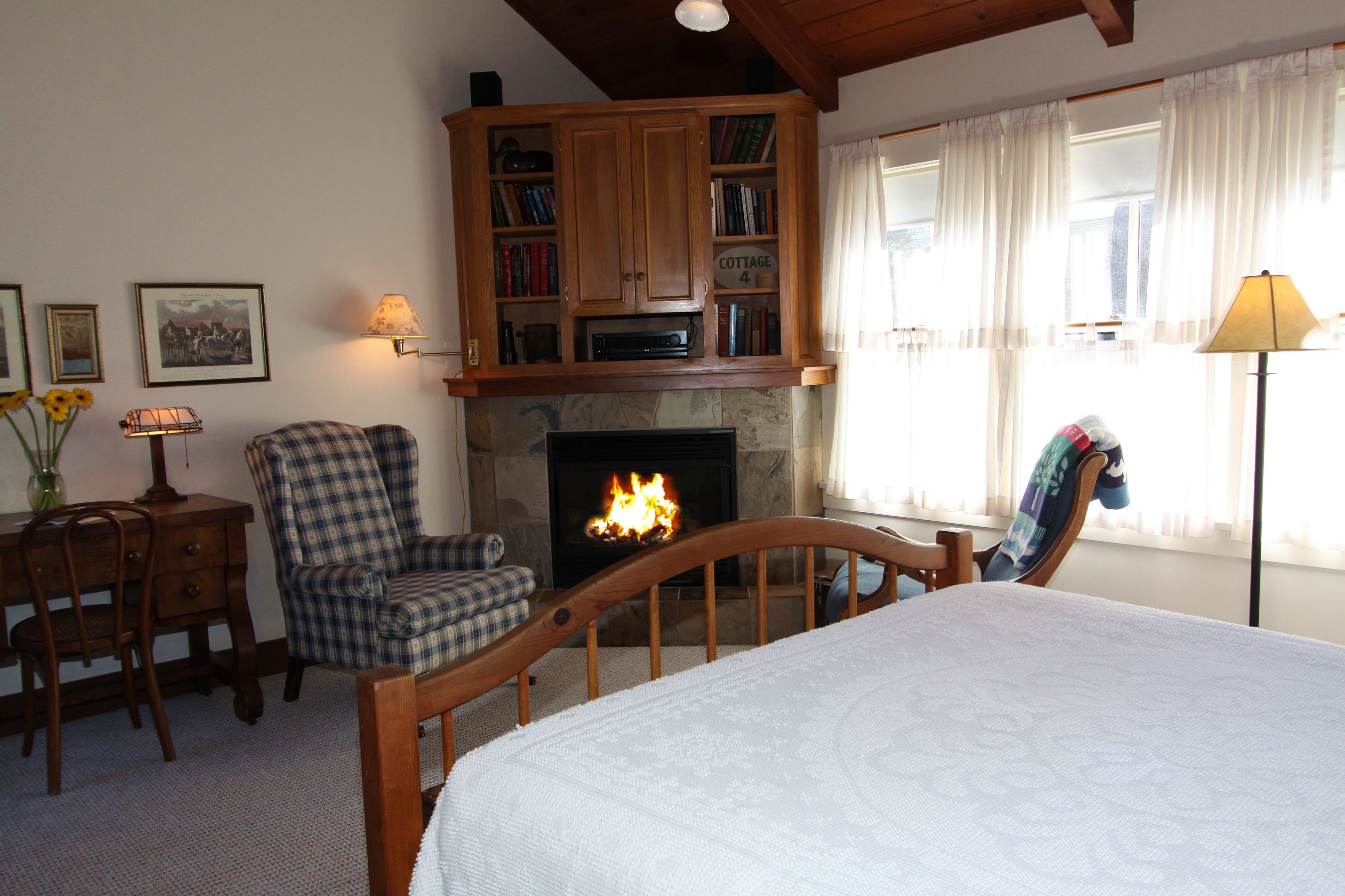 A cozy room with a fireplace, a wooden bed, a checkered armchair, a bookshelf, and a desk with a lamp.