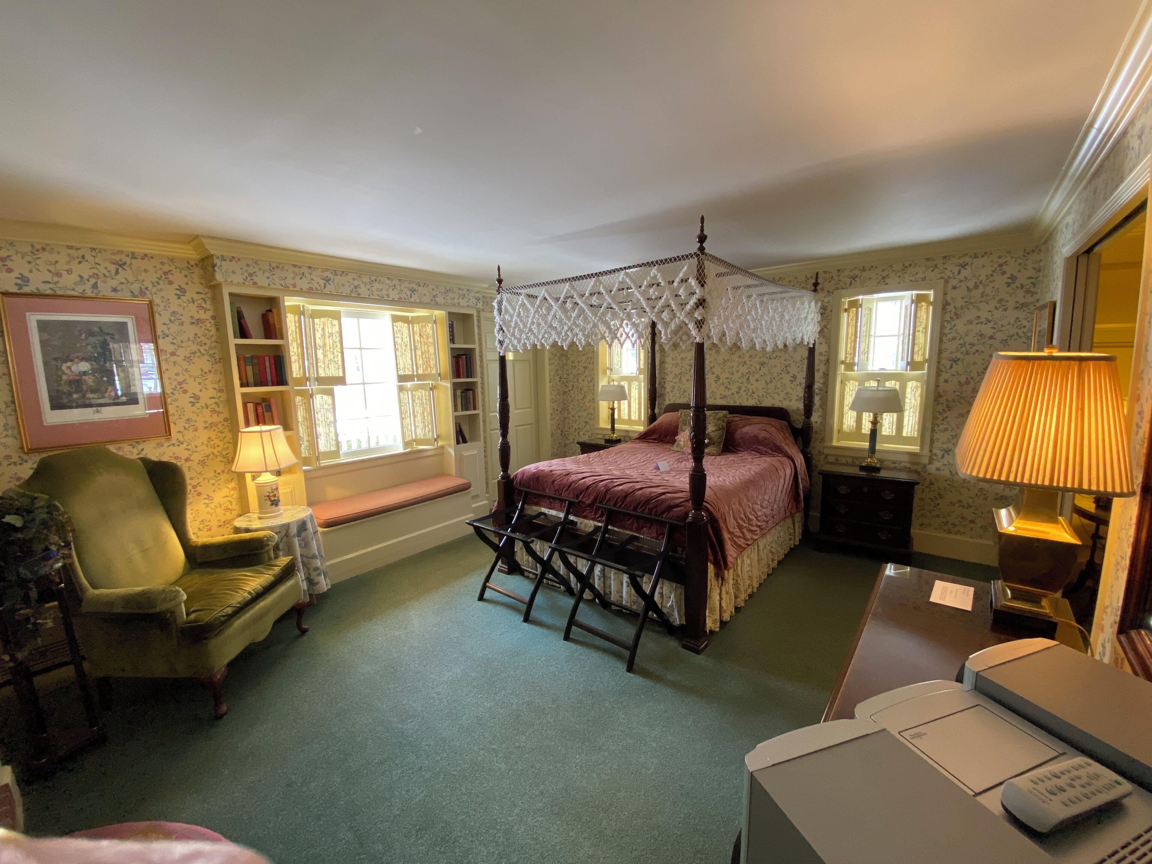 A traditional bedroom with a four-poster bed, a bedside table, a chair, a desk, and a window with a view.