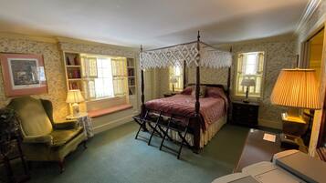 A traditional bedroom with a four-poster bed, a bedside table, a chair, a desk, and a window with a view.
