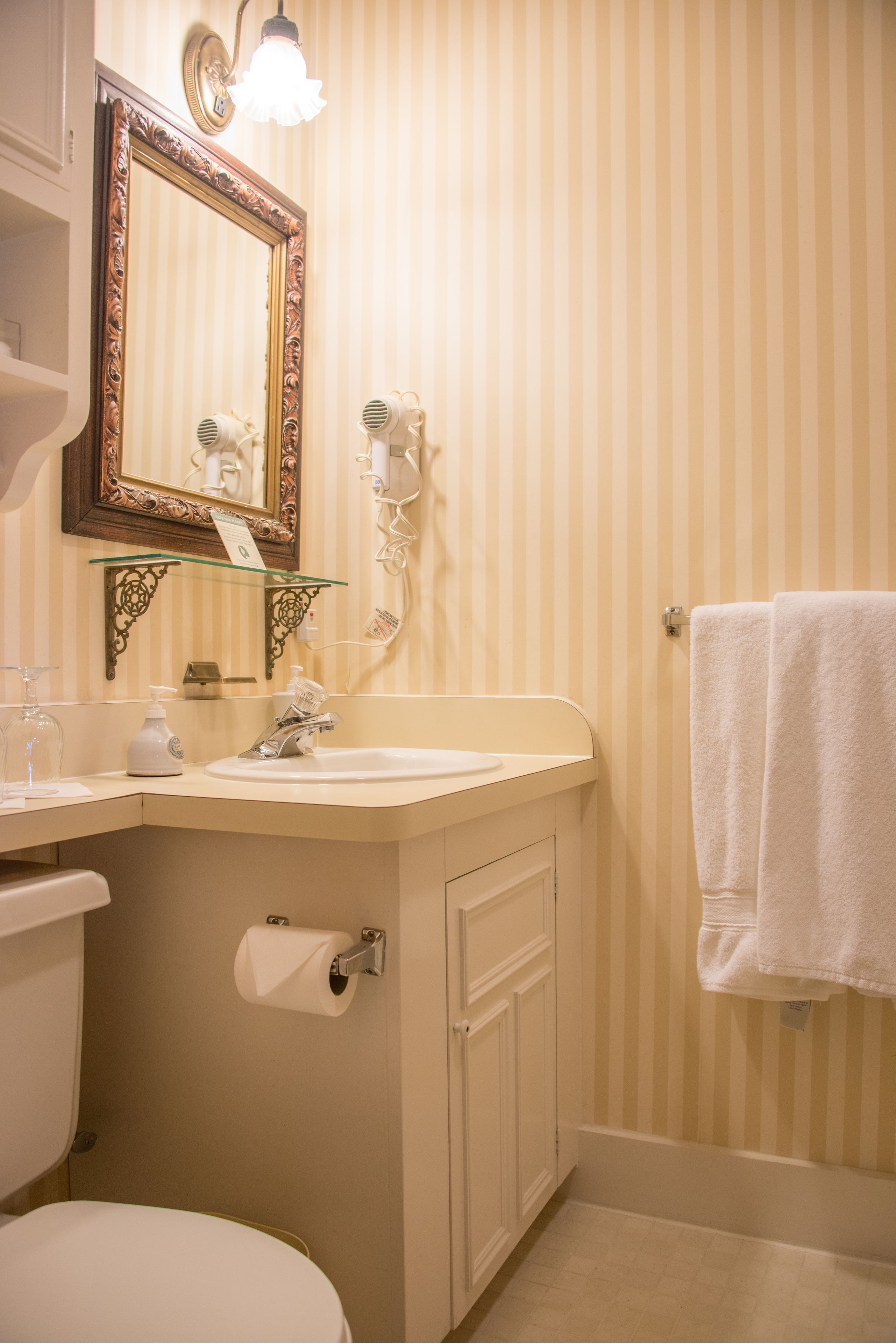 Un baño con pared a rayas, un espejo con marco decorativo, un lavabo con dispensador de jabón, una toalla colgada en la pared y un inodoro.