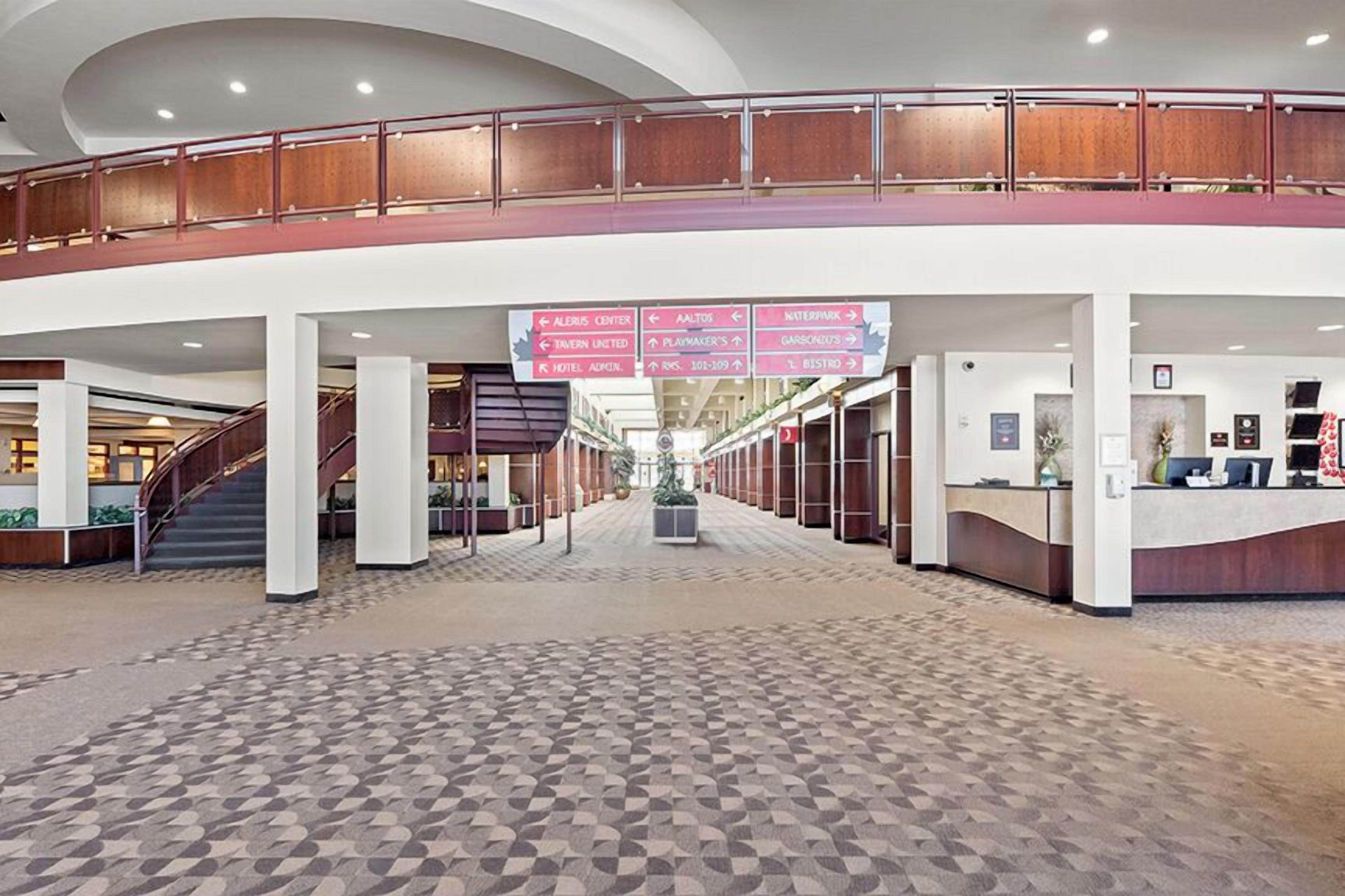 A spacious lobby with a central staircase, a reception desk, and directional signs for various hotel amenities.