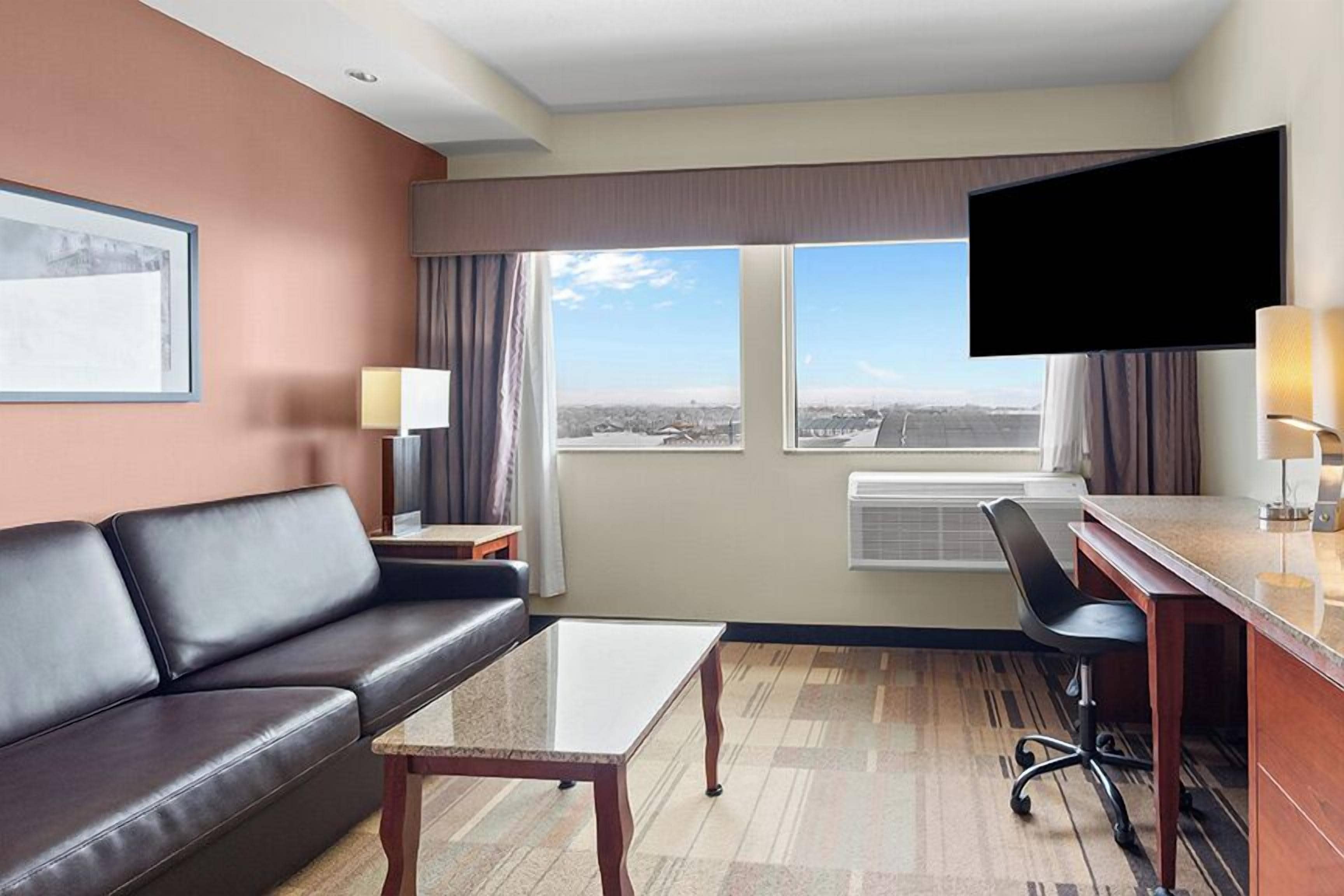 Une chambre d’hôtel équipée d’un canapé, d’une table basse, d’un bureau avec une chaise, d’un téléviseur à écran plat et d’une grande fenêtre offrant une vue dégagée.
