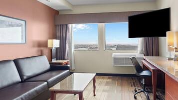 Une chambre d’hôtel équipée d’un canapé, d’une table basse, d’un bureau avec une chaise, d’un téléviseur à écran plat et d’une grande fenêtre offrant une vue dégagée.