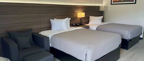 A hotel room with two beds, a desk, a chair, and a nightstand with a lamp.