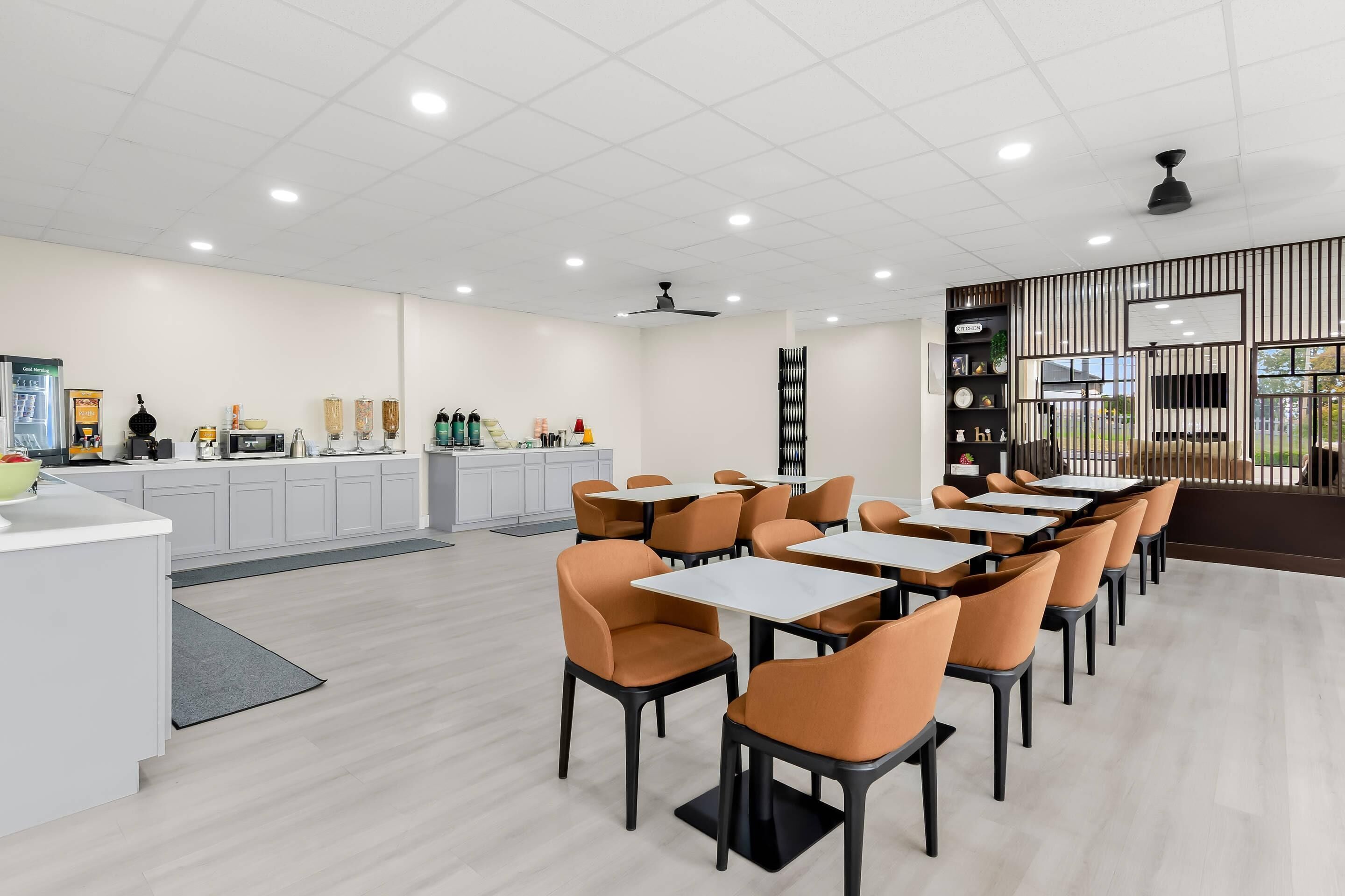 A modern dining area with white tables and orange chairs, a kitchen area with white cabinets, and a decorative wall with vertical lines.