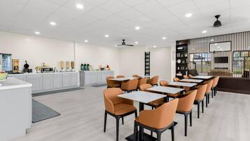 A modern dining area with white tables and orange chairs, a kitchen area with white cabinets, and a decorative wall with vertical lines.