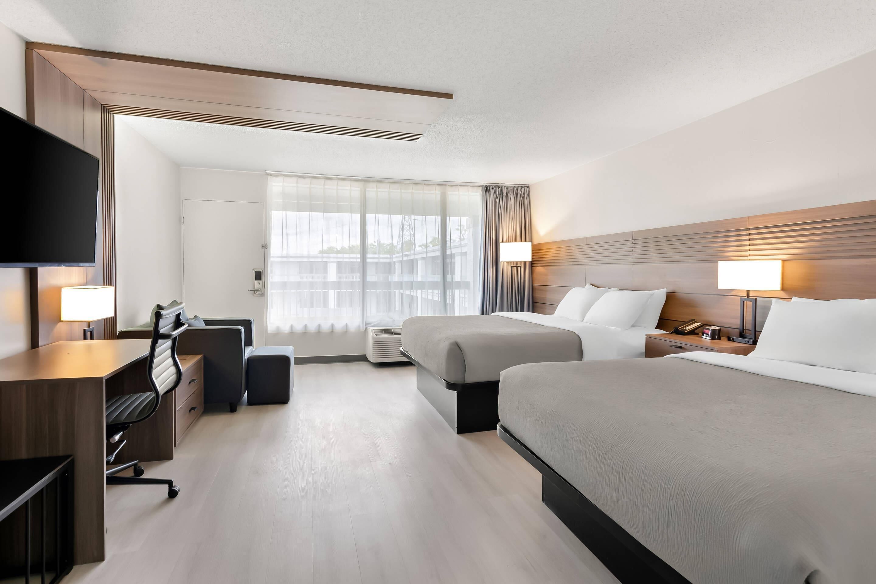 Ein Hotelzimmer mit zwei Betten, einem Schreibtisch mit Stuhl, einem Flachbildfernseher, einem Nachttisch mit Lampe und einem großen Fenster mit Vorhängen.