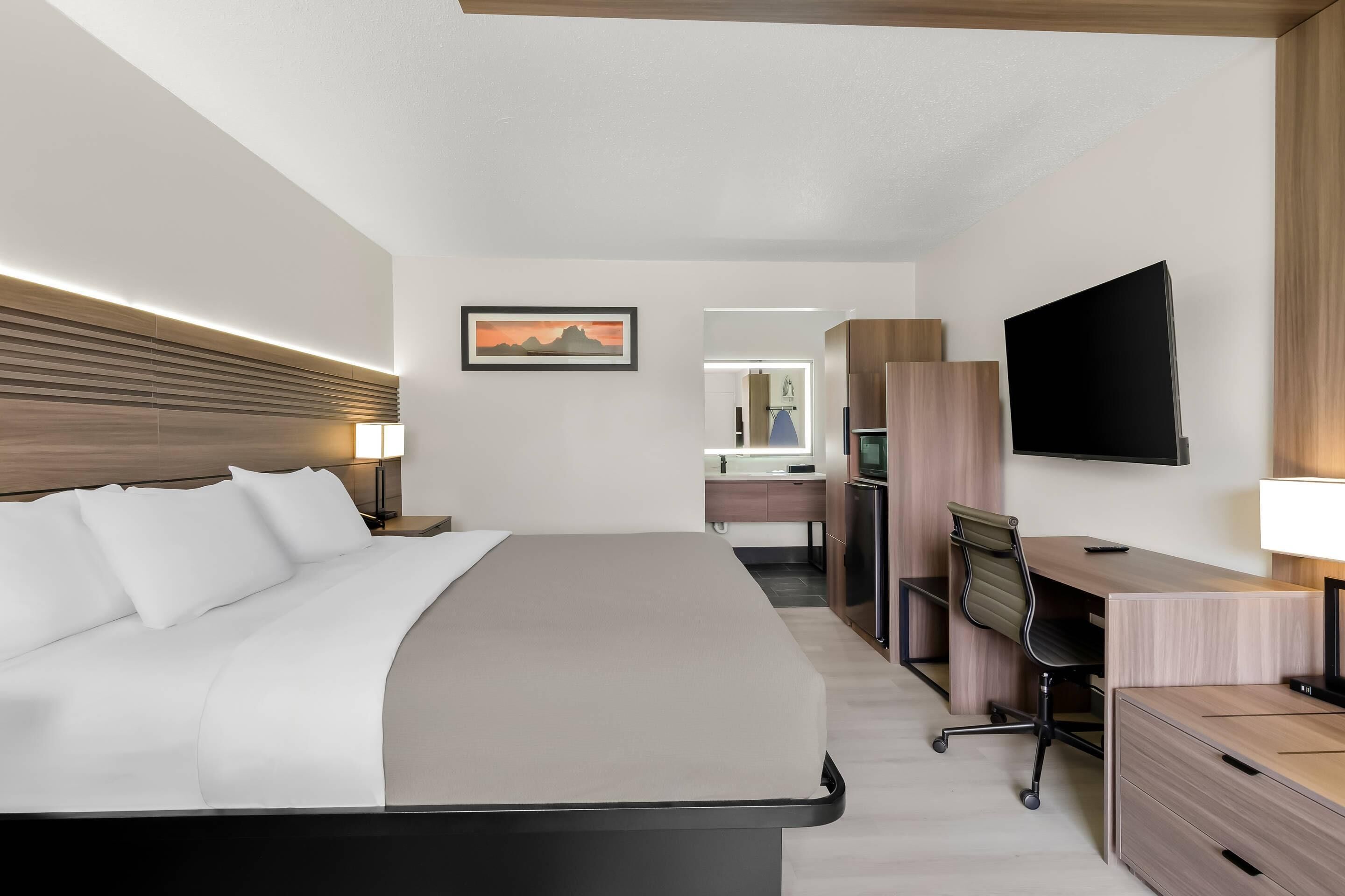 Habitación de hotel moderna con una cama grande, un escritorio, una silla, un televisor y un baño visible al fondo.