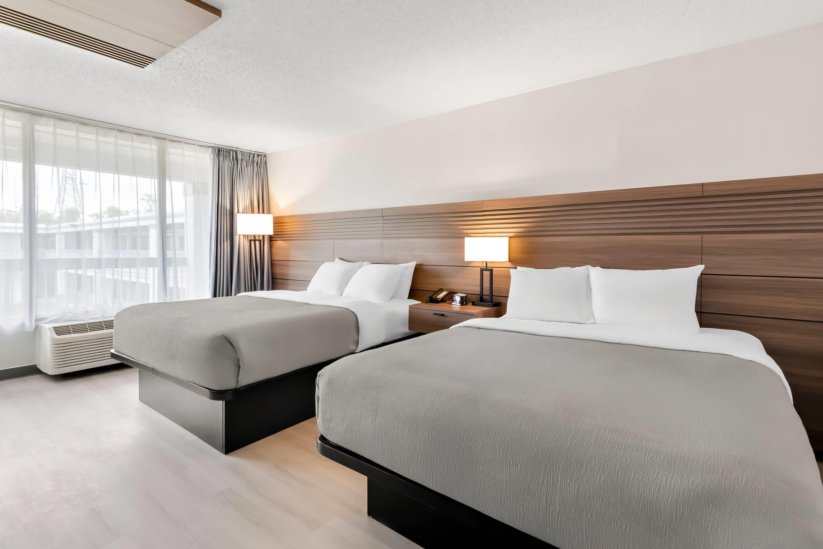 Quarto de hotel com duas camas, uma janela grande com cortinas, uma mesa de cabeceira com abajur e um aquecedor fixado na parede.