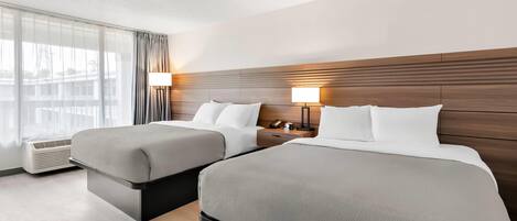 Una habitación de hotel con dos camas, un ventanal con cortinas, una mesita de noche con lámpara y un calentador de pared.
