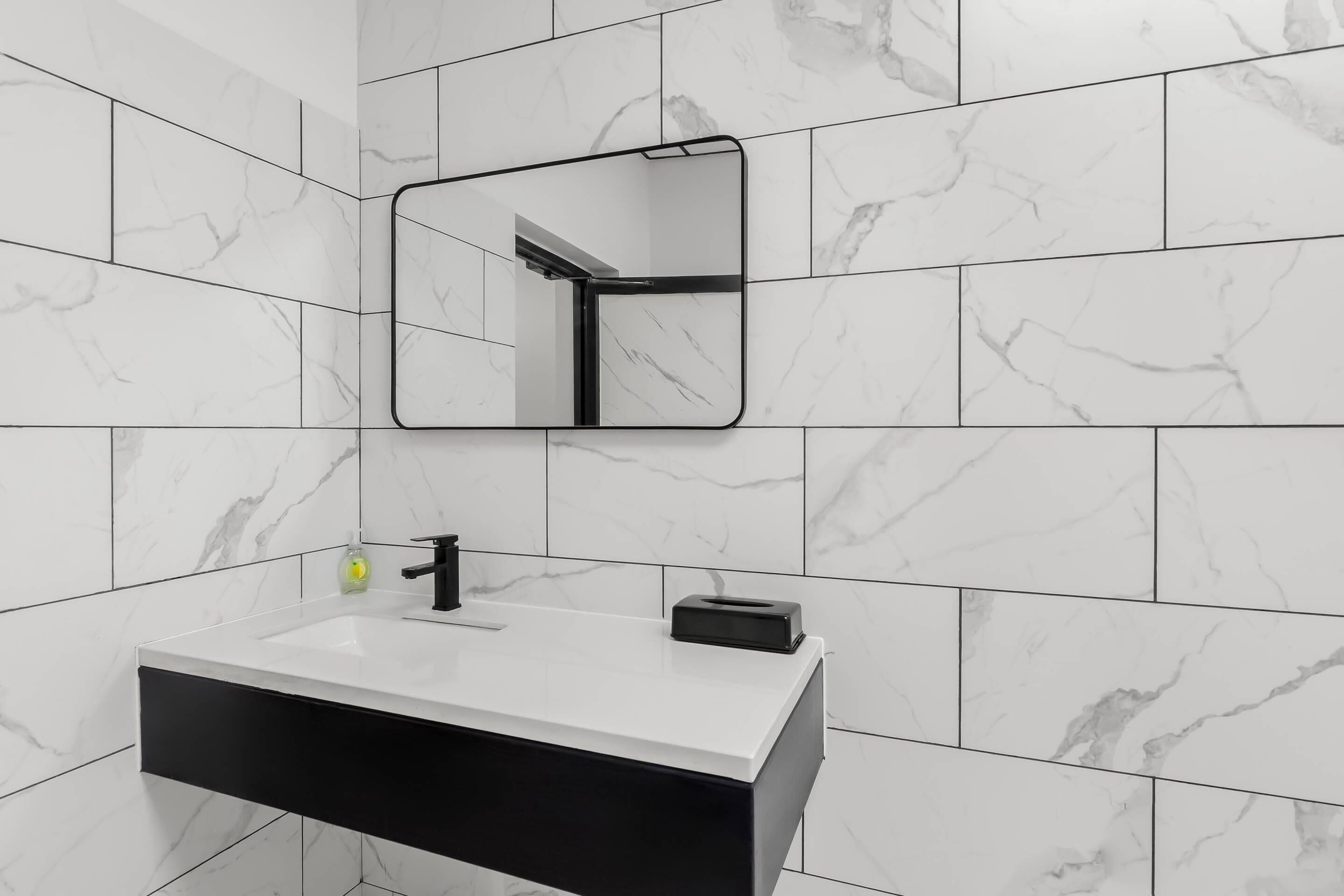 A modern bathroom with a white marble tiled wall, a black and white vanity, a mirror, and a soap dispenser.