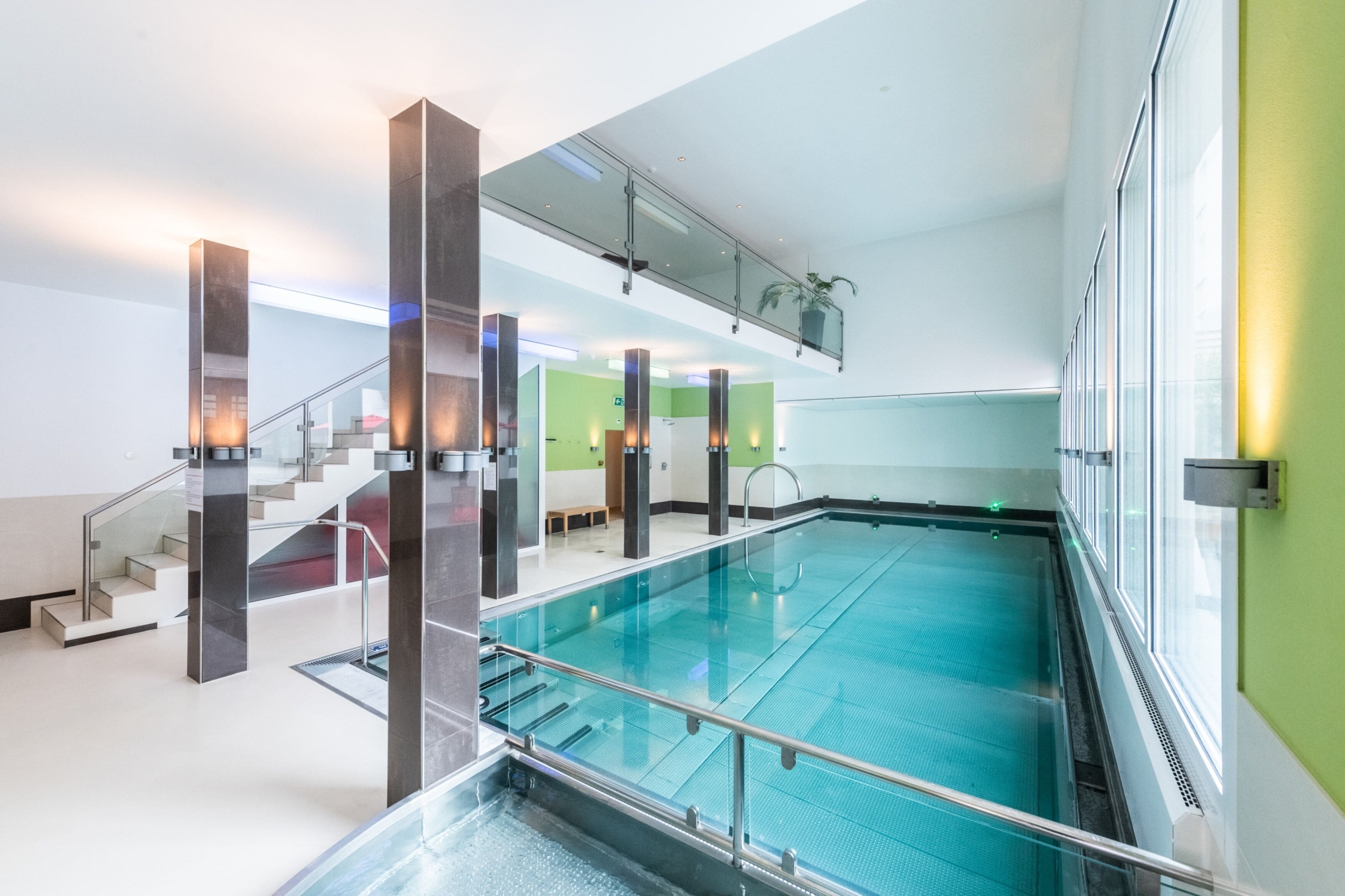 Ein moderner, überdachter Schwimmbereich mit Glasgeländern, einer Treppe mit Metallgeländern und einer grünen Wand im Hintergrund.