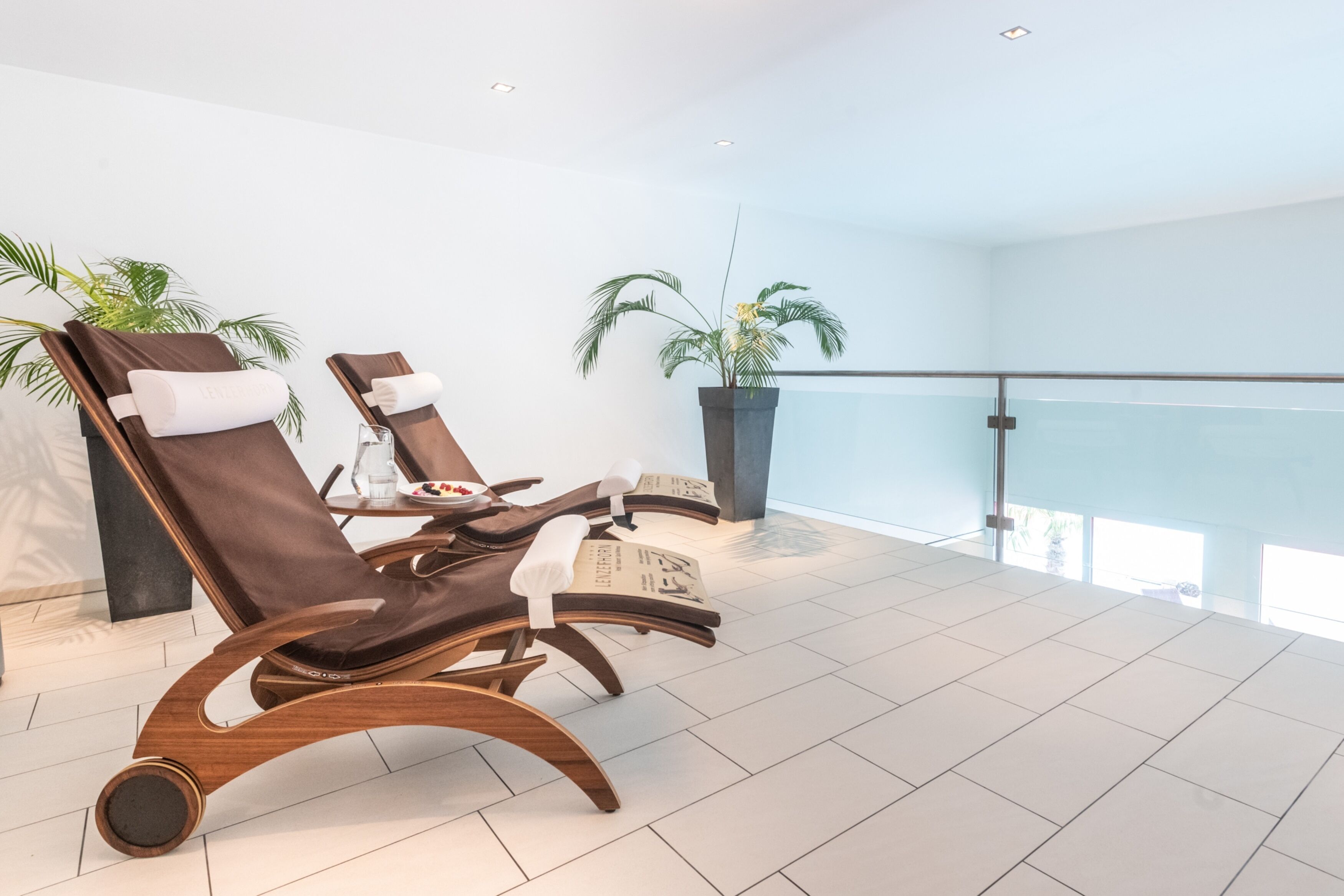 A modern lounge area with two wooden lounge chairs, a potted plant, and a glass balustrade overlooking a balcony.