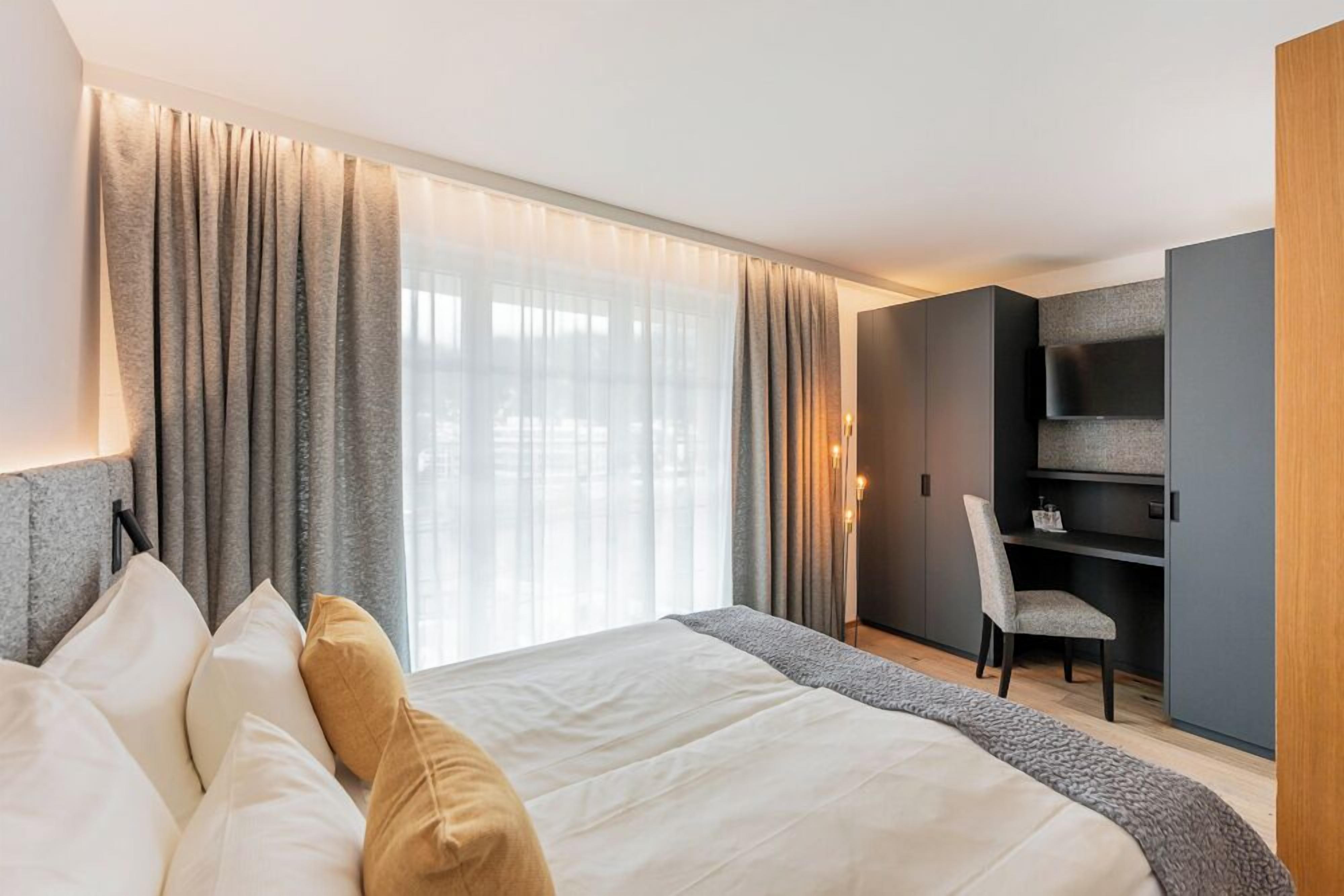 A modern hotel room with a large bed, a desk with a chair, a TV, and a large window with curtains.