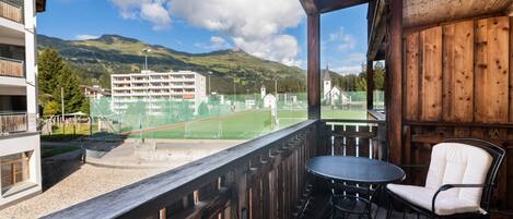 Balcón de madera con una mesa y una silla, con vista a una cancha de tenis y edificios.