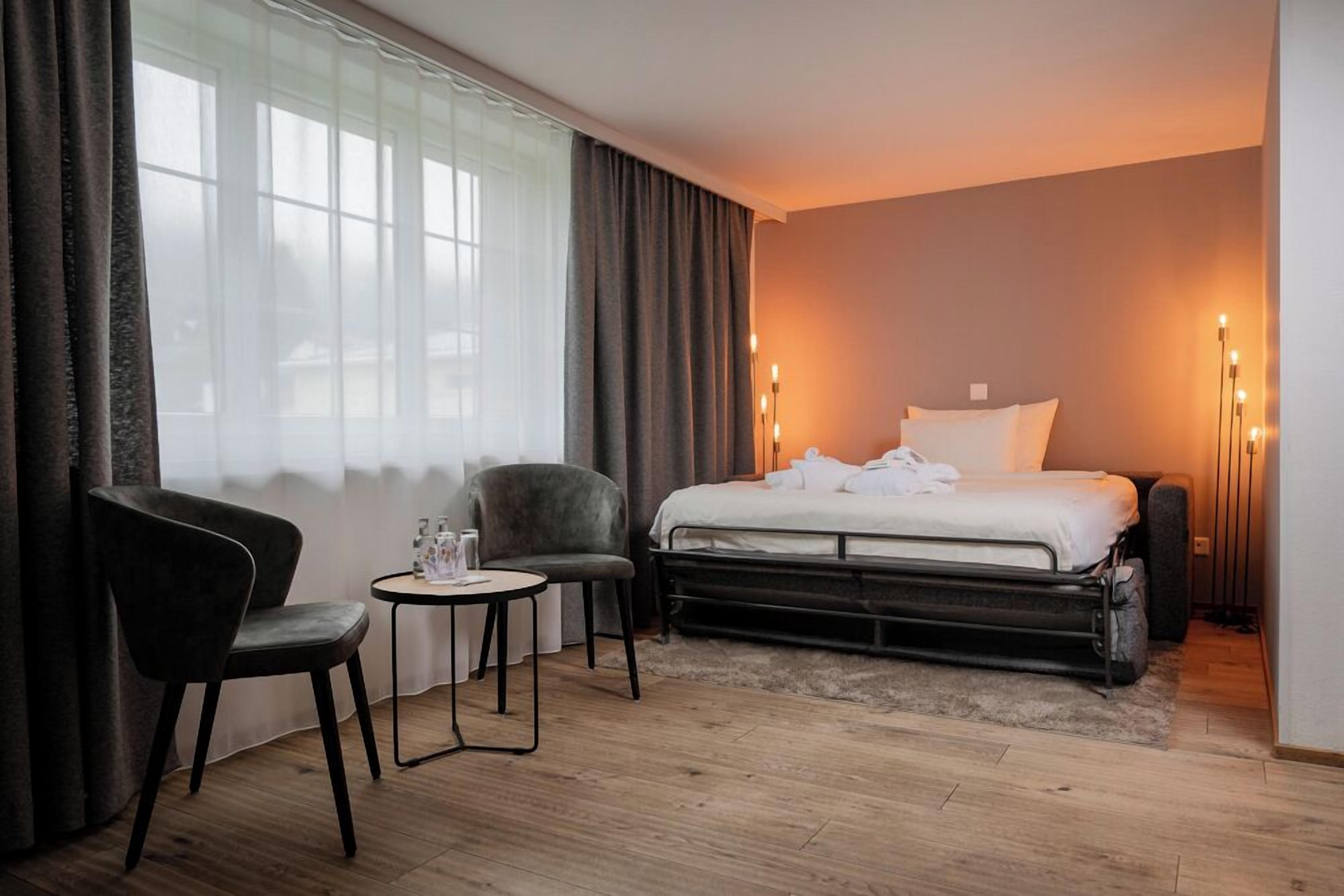 A hotel room with a bed, two chairs, a small table, and a bedside lamp.