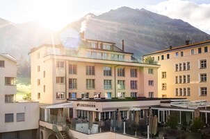 Un hotel con terraza, un restaurante llamado "LENZERHORN" y un paisaje de montañas al fondo.