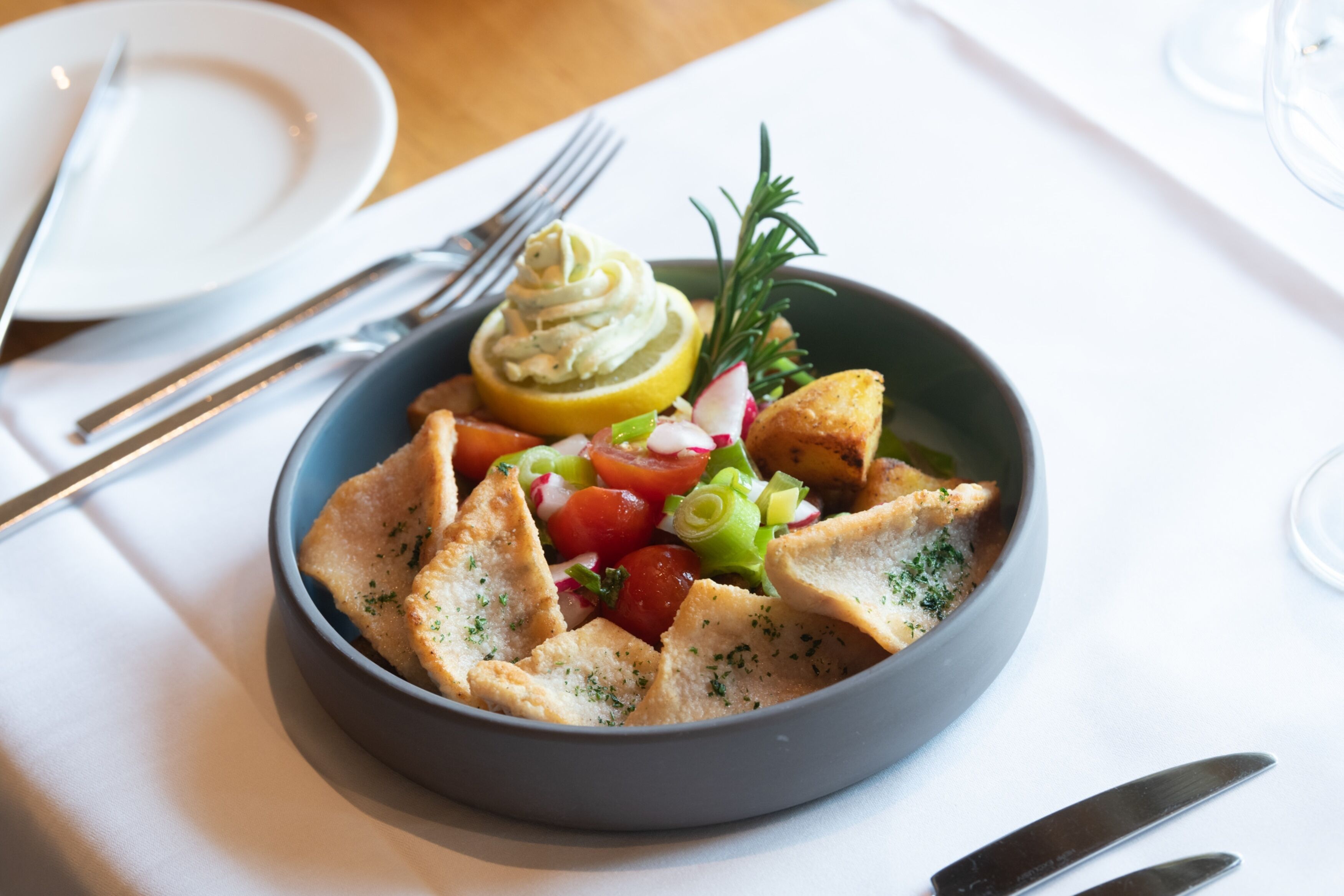 Une salade servie dans un bol, accompagnée d’une tranche de citron, de radis et d’oignons verts, ainsi que de croûtons et d’une cuillère de crème.