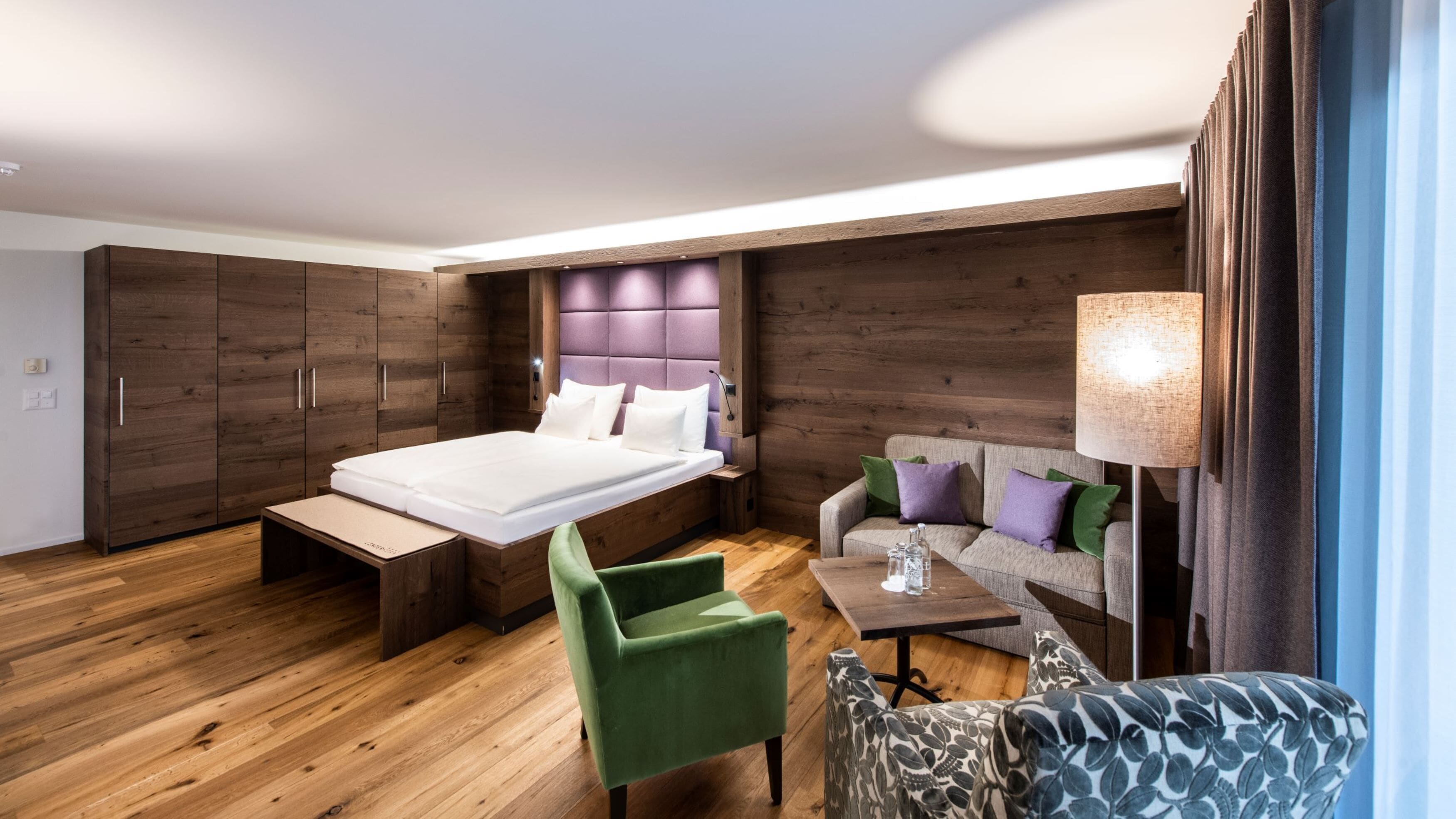 Una habitación de hotel moderna con una cama grande, un armario de madera, un sillón verde, un sofá con cojines estampados y una mesita con una lámpara.