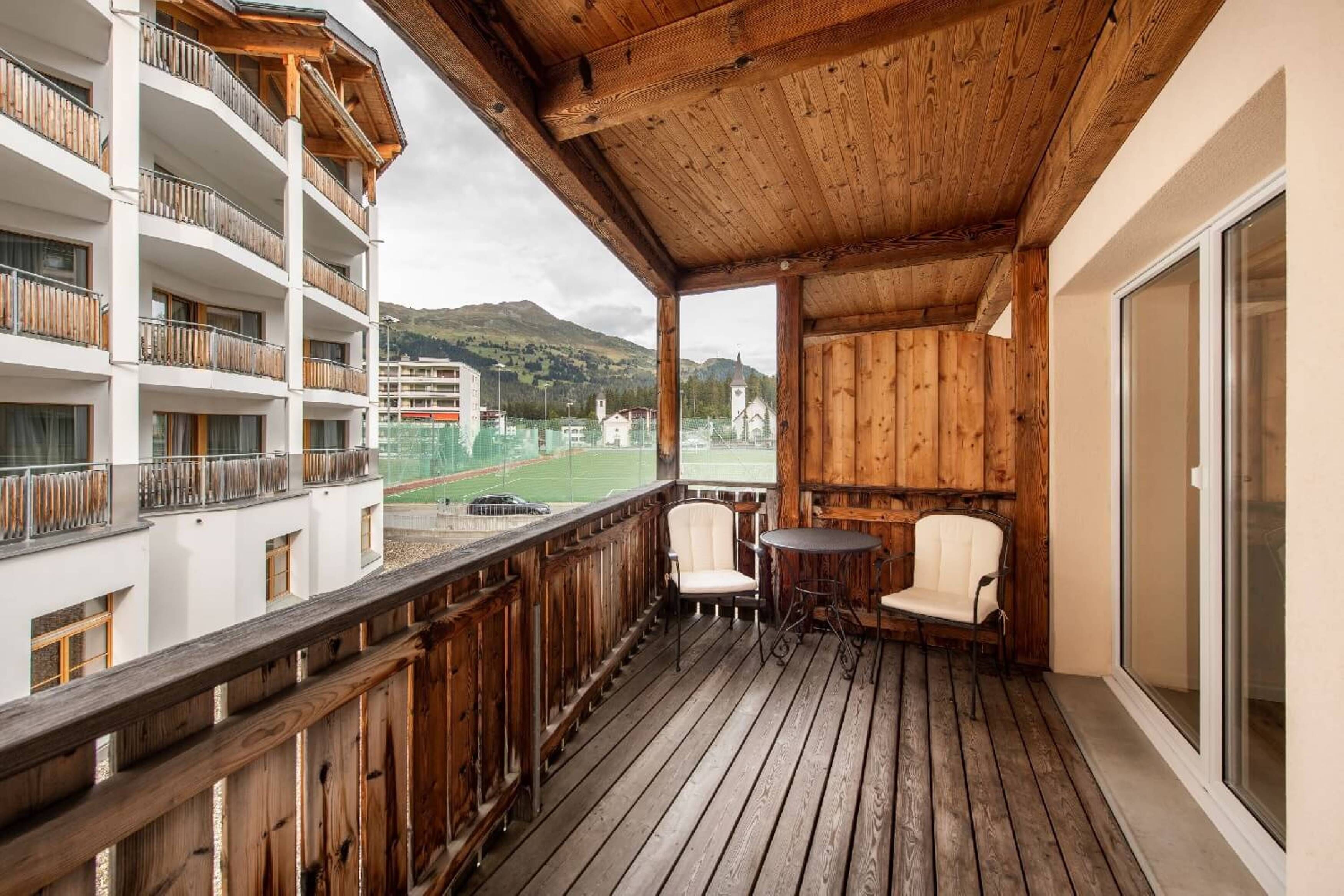 Ein Holzbalkon mit zwei Stühlen und einem Tisch, mit Blick auf ein Sportfeld und Gebäude.
