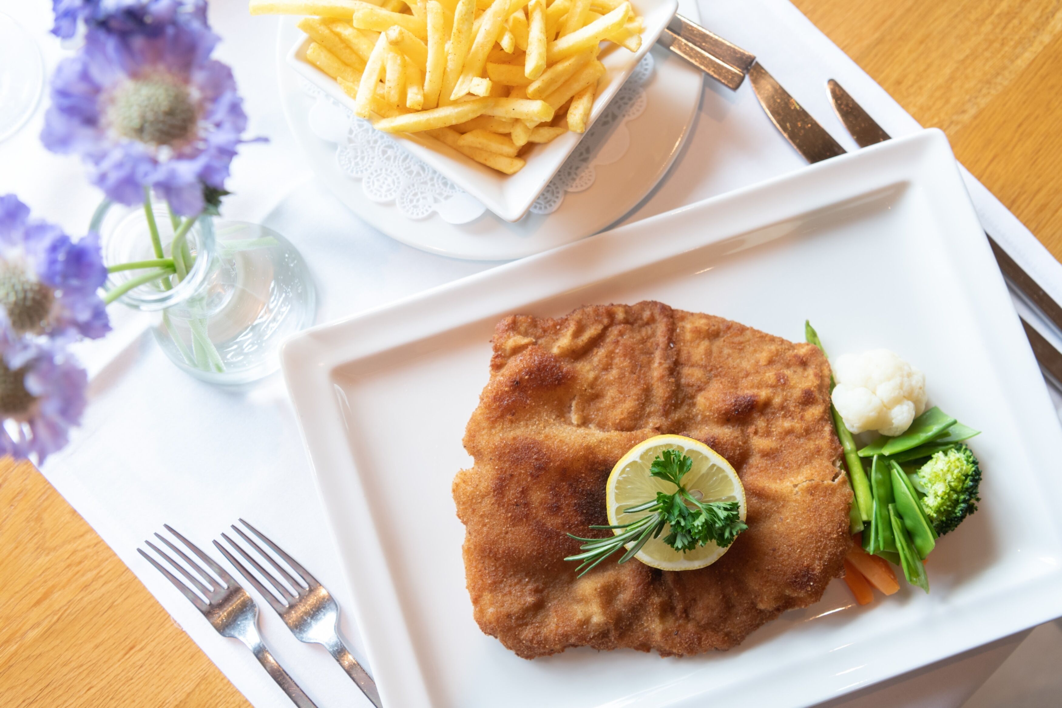 Un plato con un filete empanizado, limón, perejil y verduras, acompañado de un bol de papas fritas.