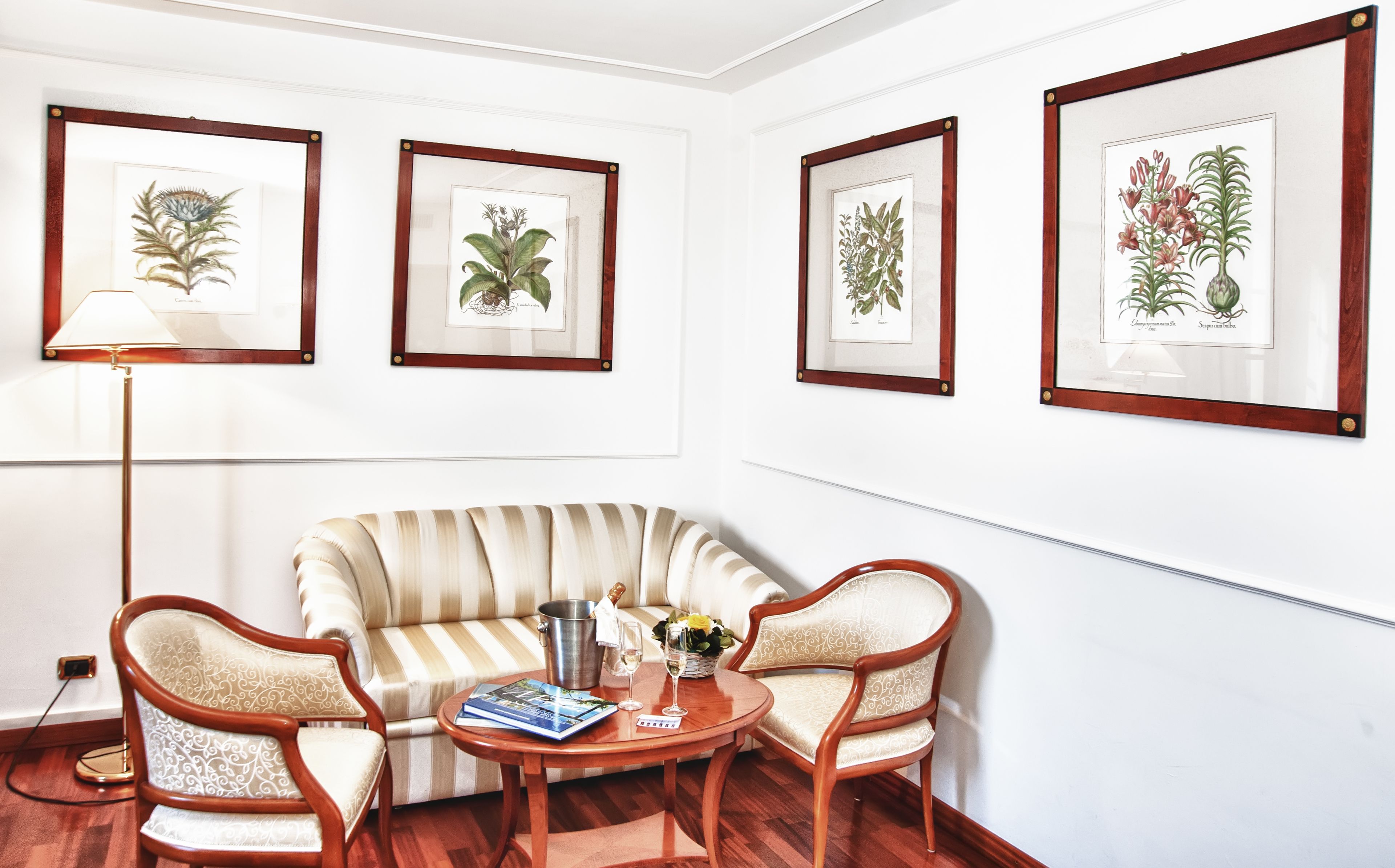 A living room with a striped sofa, two wooden chairs, a small round table with a book and a vase of flowers, and framed botanical illustrations on the wall.