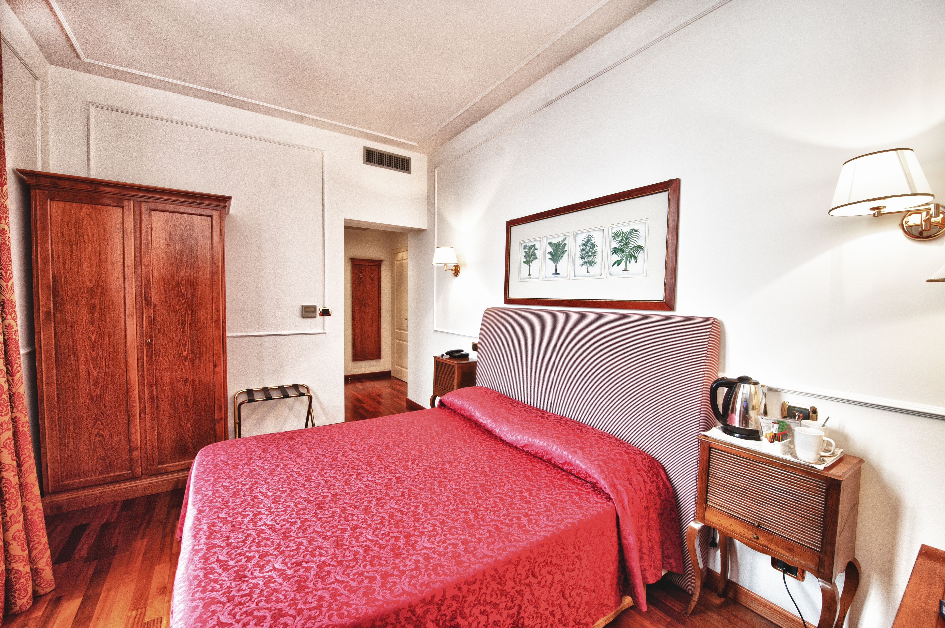 A hotel room with a bed, a wooden wardrobe, a framed picture on the wall, a bedside table with a lamp, and a radiator.