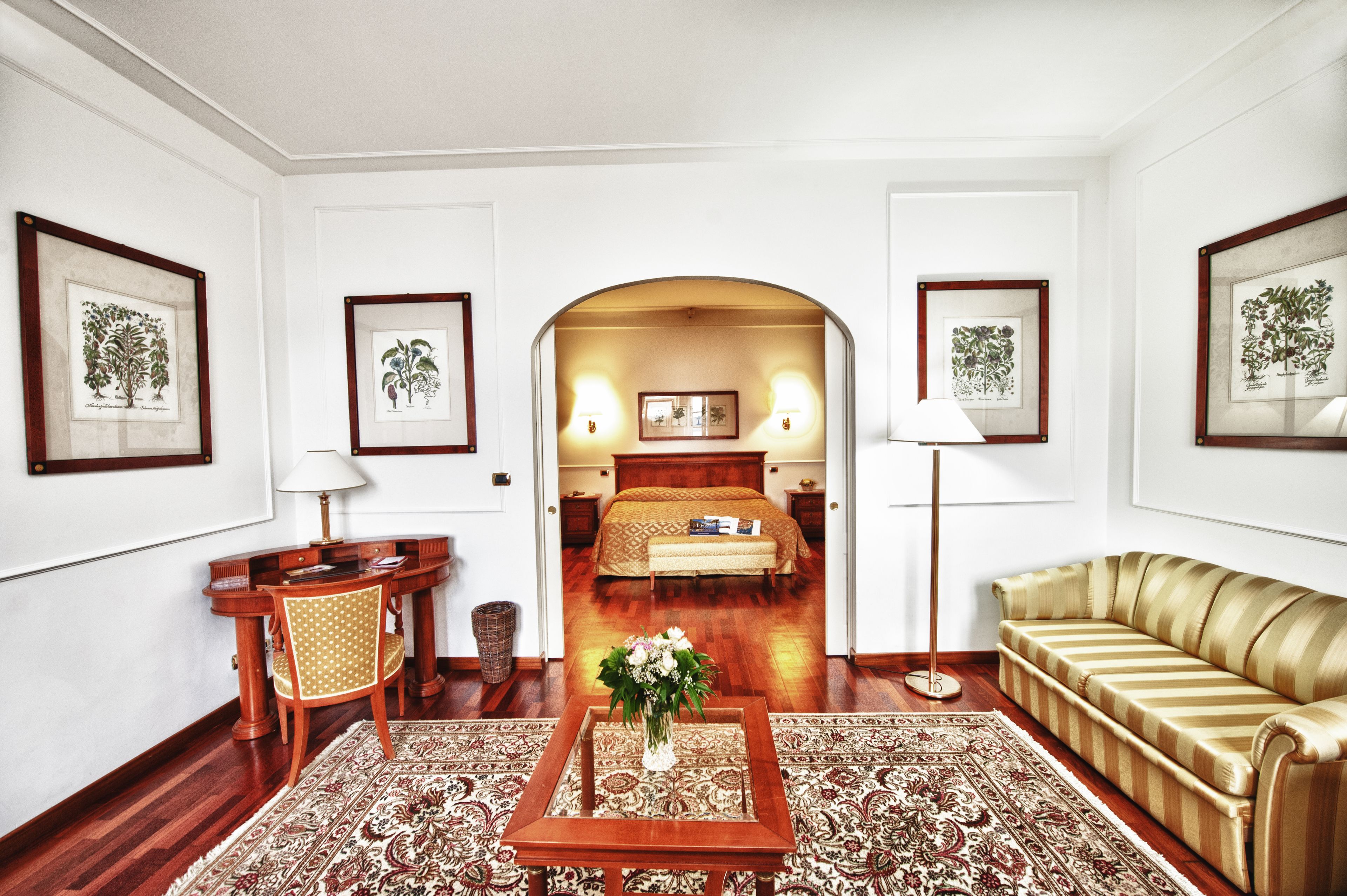 A hotel room with a wooden floor, a sofa, a dining table with chairs, a coffee table with a vase, and framed botanical prints on the walls.