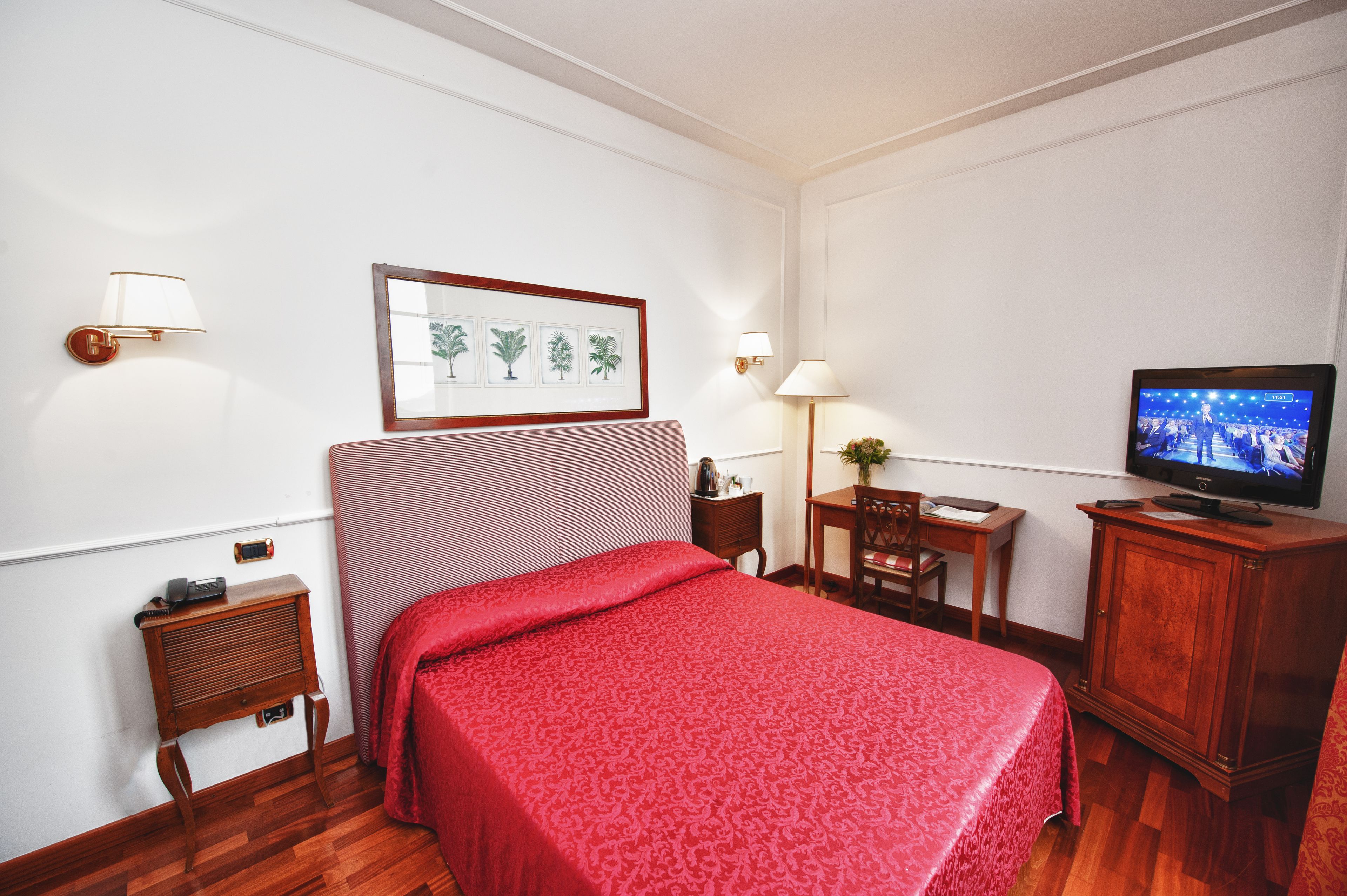 A hotel room with a bed, a television, a nightstand, a lamp, a framed picture, and a small table.