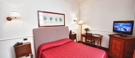 A hotel room with a bed, a television, a nightstand, a lamp, a framed picture, and a small table.