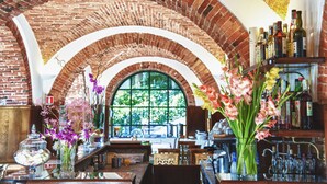 Zona de bar con techo de ladrillo, ventanas arqueadas y varias botellas y flores sobre estantes.