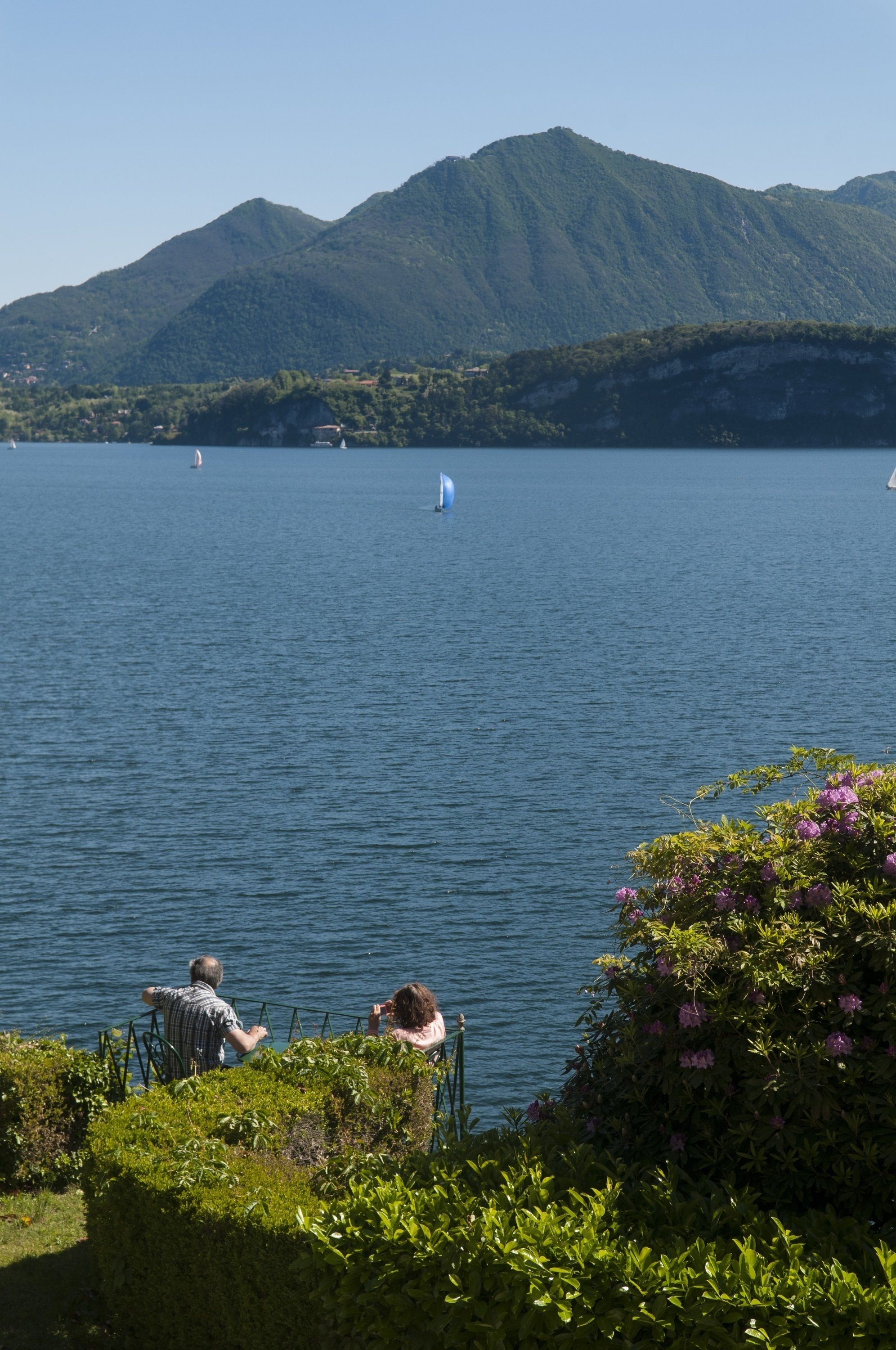 A lake with a mountain in the background, a sailboat, and two people sitting on a bench.