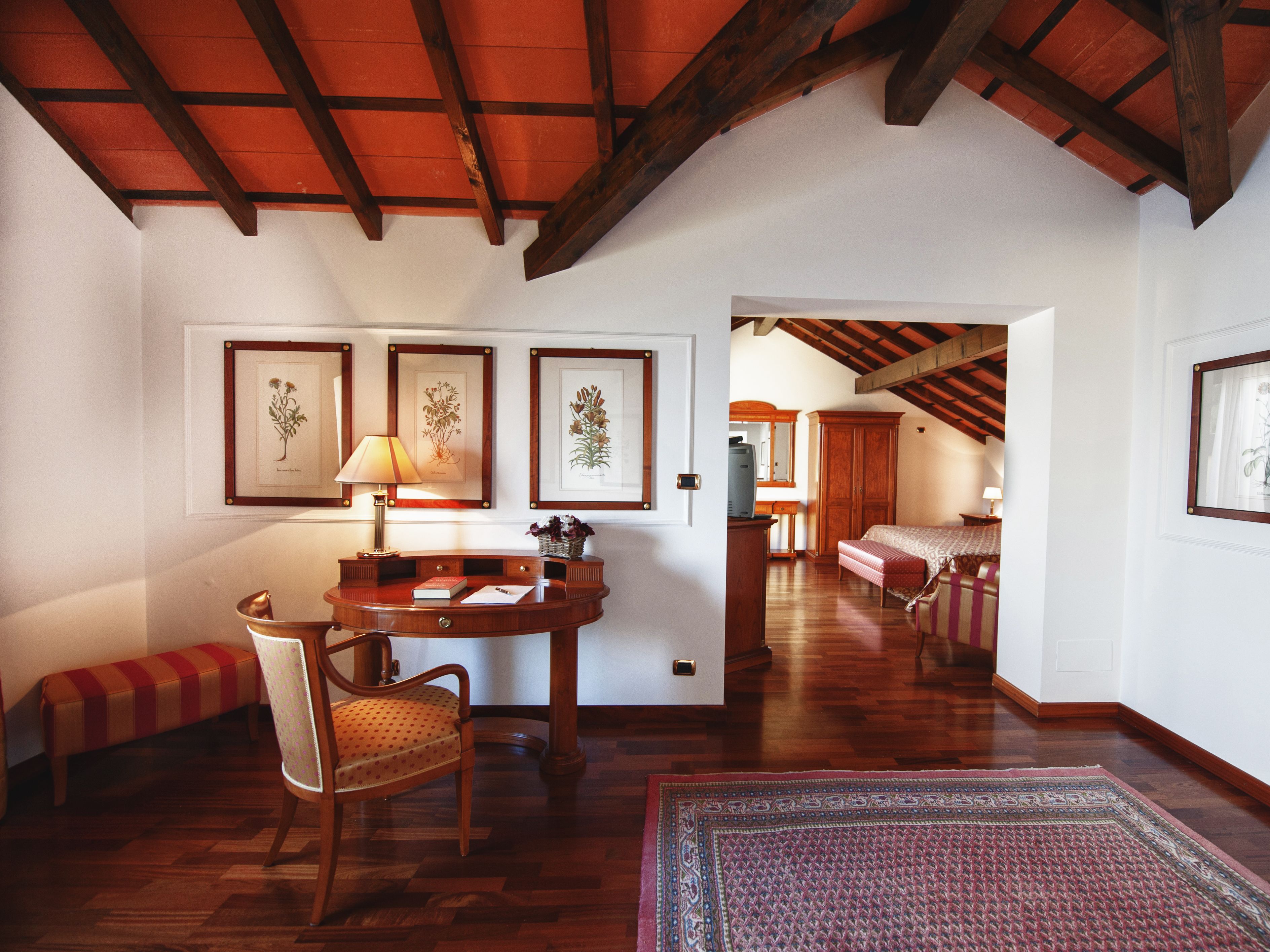 A room with wooden beams, a desk with a lamp, a chair, a bench, and framed botanical prints on the wall.
