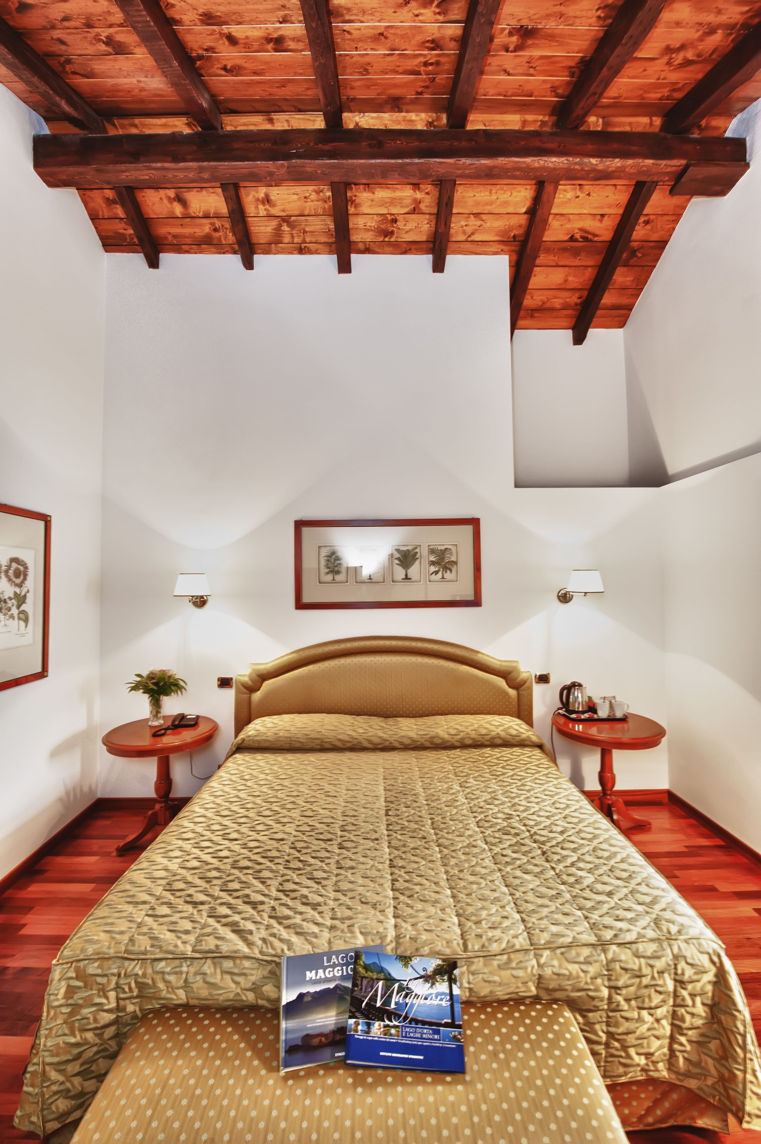 A bedroom with a wooden ceiling, a bed with a patterned bedspread, two bedside tables with lamps, and framed pictures on the walls.