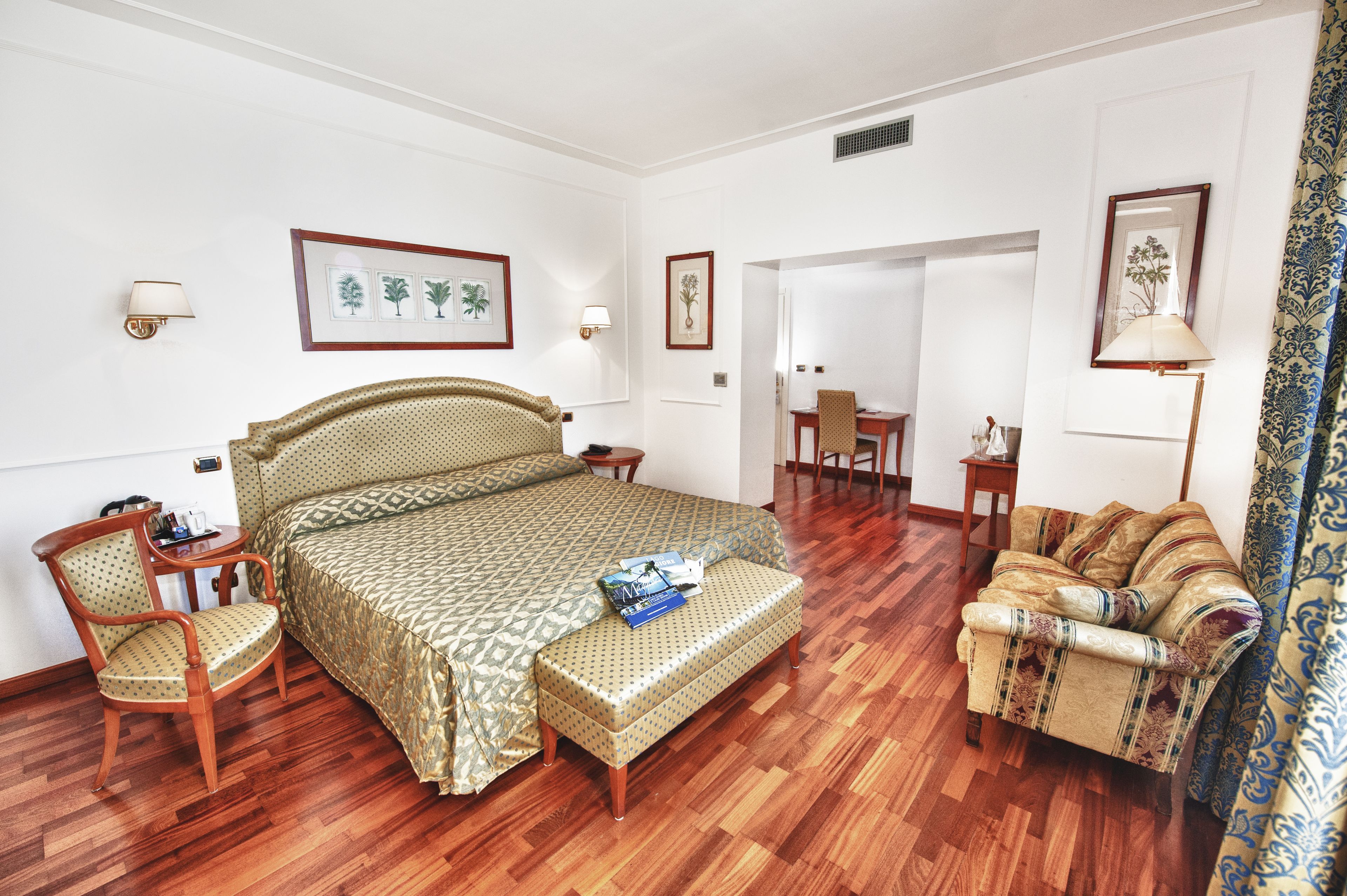 Quarto de hotel com uma cama grande, uma cadeira de madeira, um sofá e uma escrivaninha. Há quadros nas paredes e uma luminária na escrivaninha.