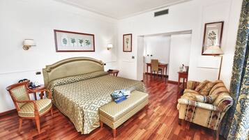 Quarto de hotel com uma cama grande, uma cadeira de madeira, um sofá e uma escrivaninha. Há quadros nas paredes e uma luminária na escrivaninha.