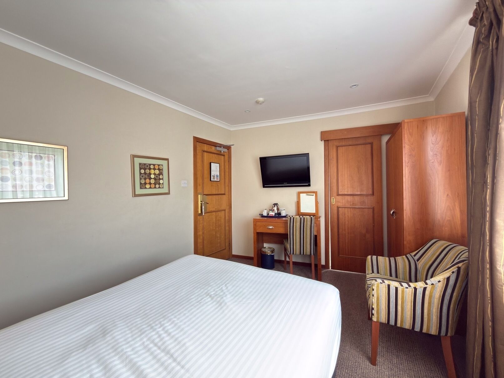A hotel room with a bed, a television, a desk with a chair, and a wardrobe.