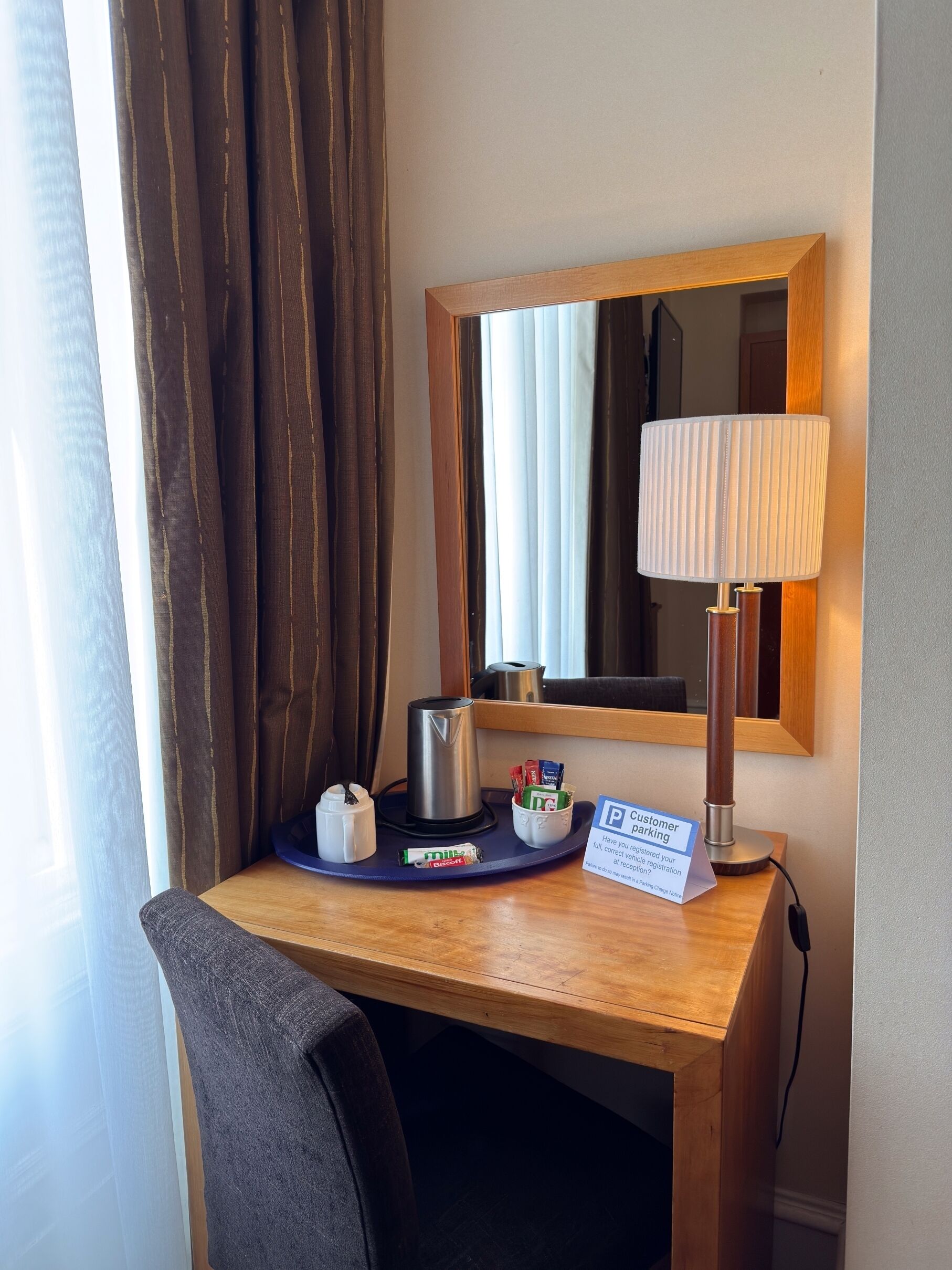 A hotel room with a wooden desk, a mirror, a lamp, a coffee maker, a tissue box, and a sign for customer parking.