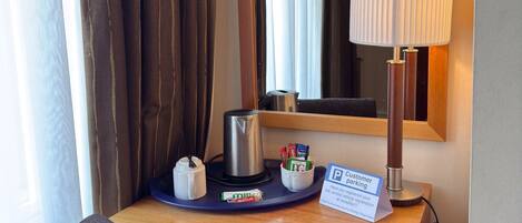 A hotel room with a wooden desk, a mirror, a lamp, a coffee maker, a tissue box, and a sign for customer parking.