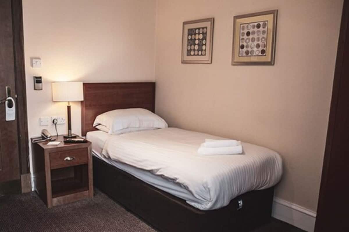 A hotel room with a single bed, a bedside table with a lamp, and two framed pictures on the wall.