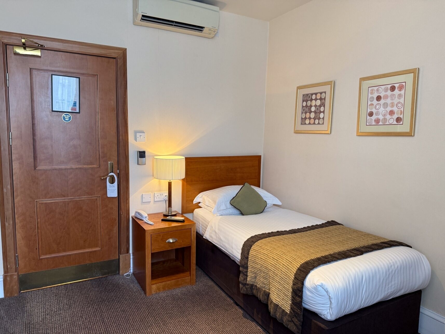 A hotel room with a wooden door, a bedside table with a lamp, a bed with a headboard, and two framed pictures on the wall.
