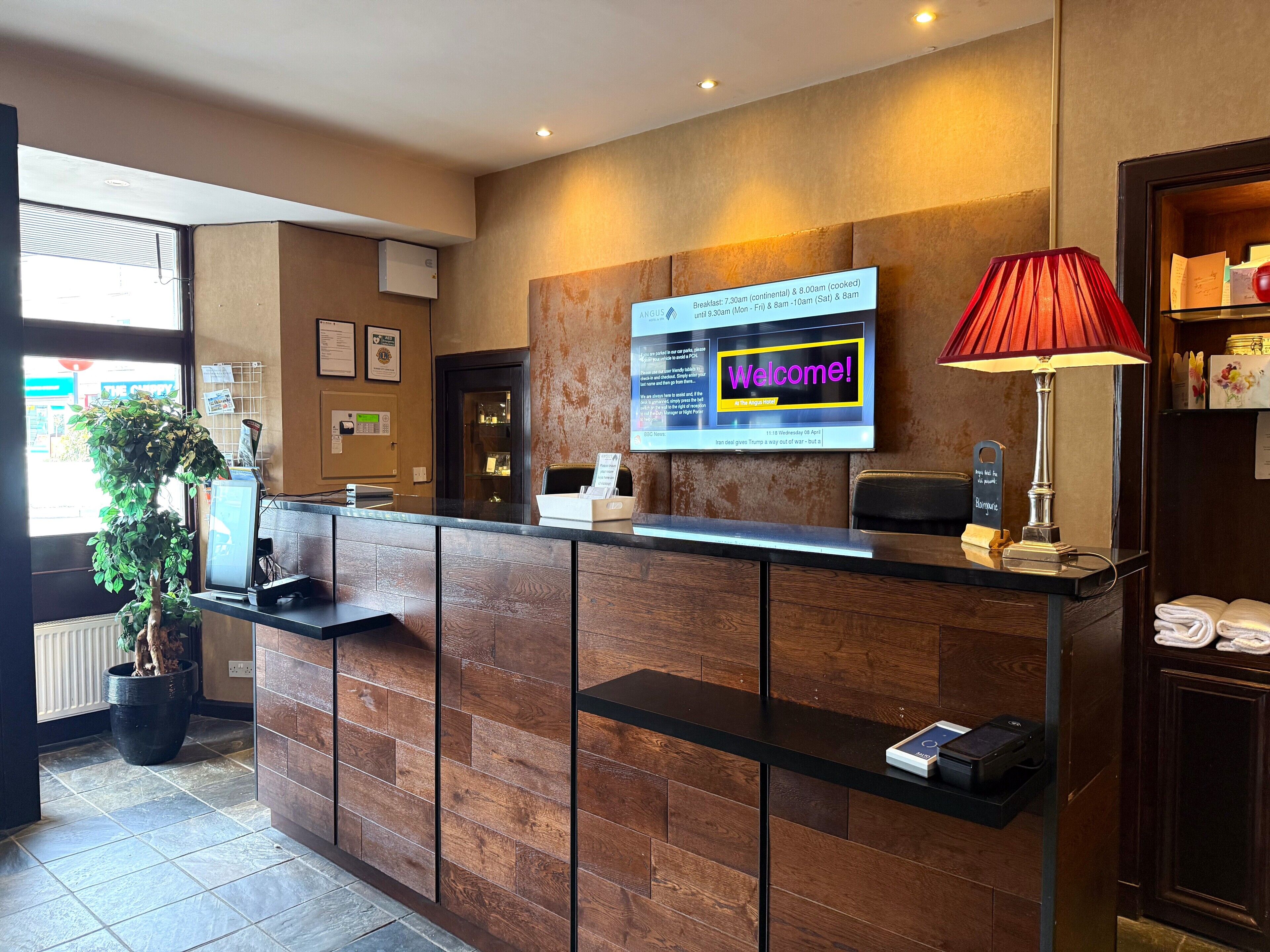 A reception desk with a digital welcome sign, a red lamp, and a potted plant.